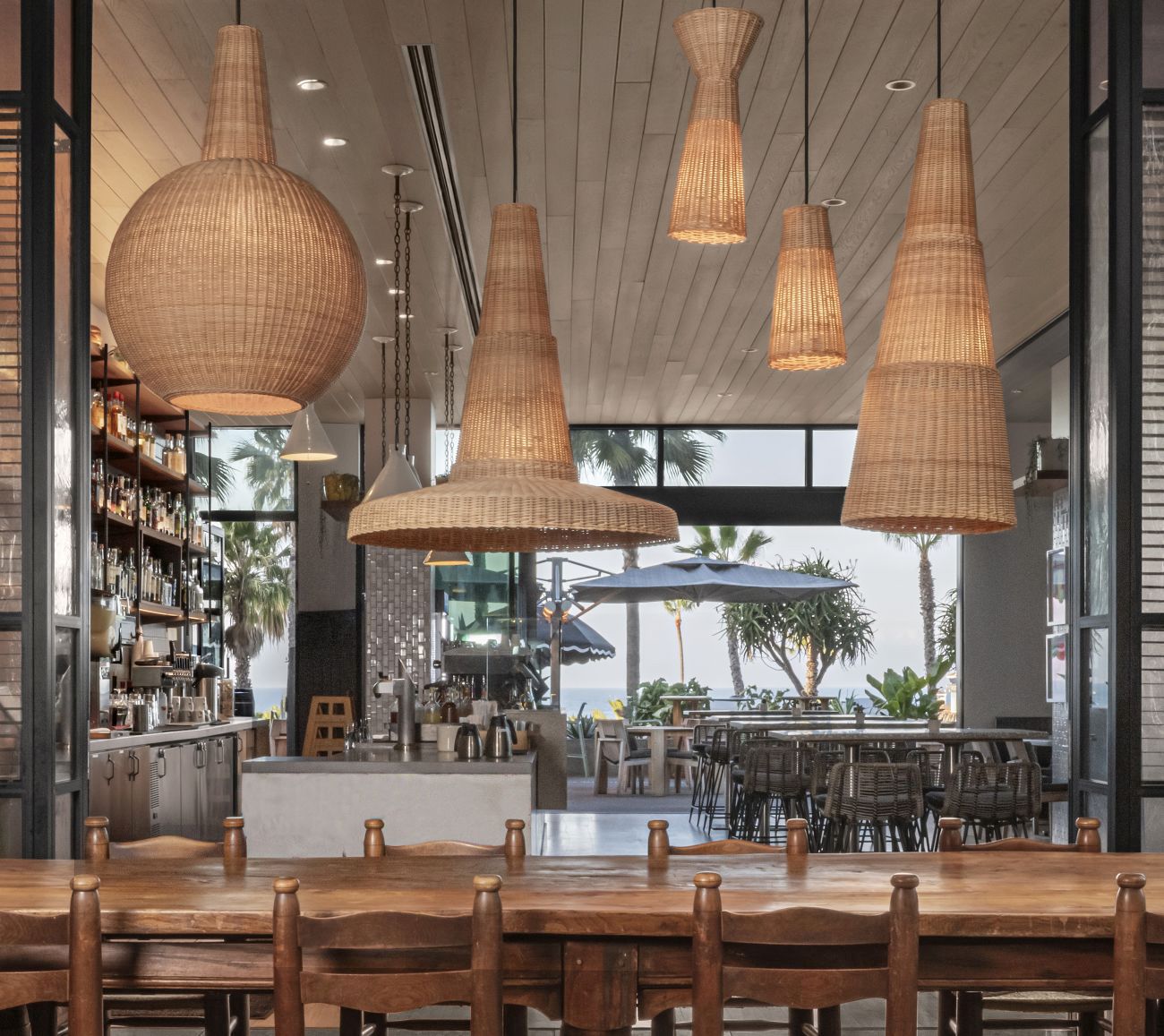 Modern open-air restaurant interior with woven pendant lights, wooden tables, and ocean-view patio seating.