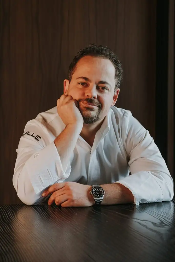 Michelin-starred chef at Valle restaurant seated at a wooden table, wearing a white shirt and watch.