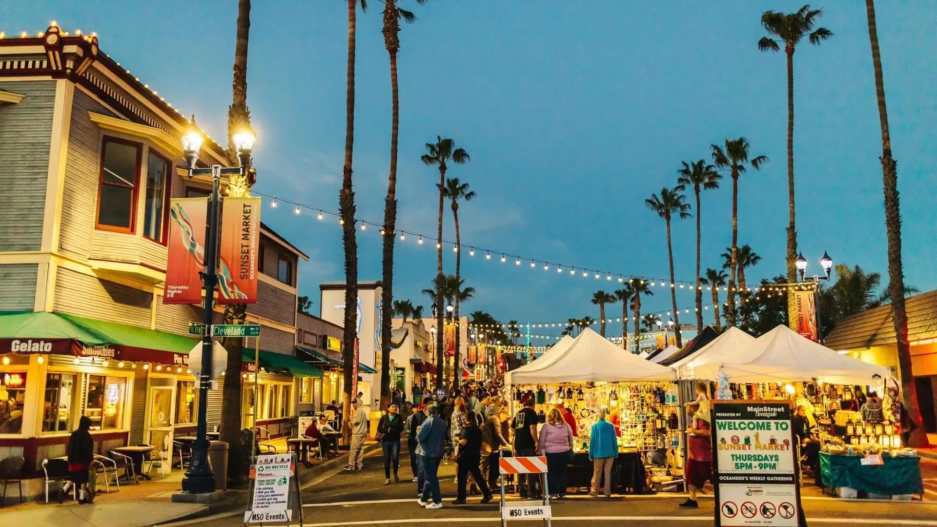 Evening street market scene at Sunset Market in Oceanside with palm trees, string lights, vendor tents & crowds enjoying food