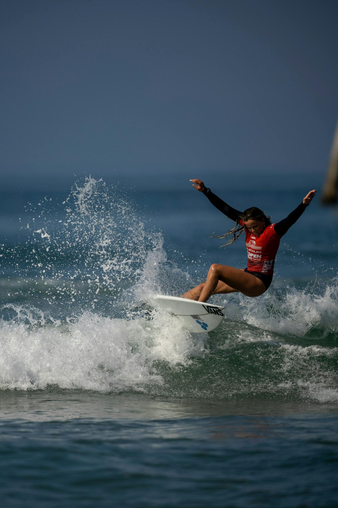 Surfer performing a dynamic maneuver on a breaking wave, wearing a red competition jersey, with water spraying around the board against a deep blue ocean backdrop.